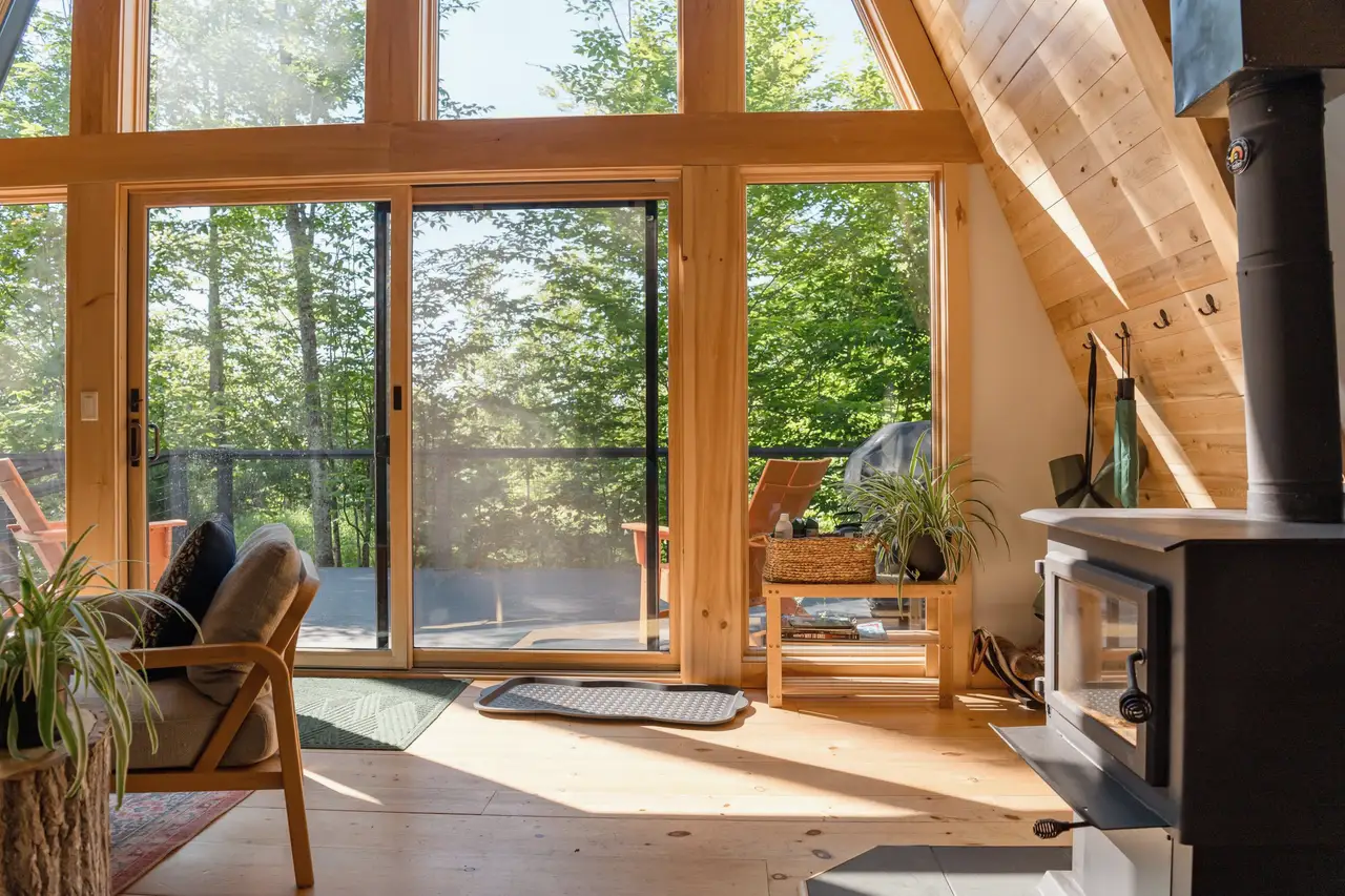 Sunlit A-frame cabin with tall windows and a sliding door onto a deck among evergreens